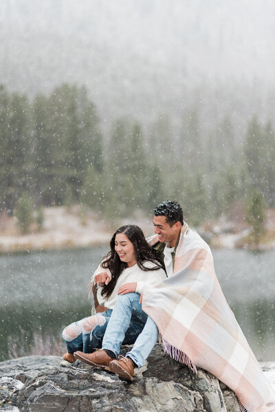 man and woman sitting on a rock together with a flannel blanket around them as they cuddle while the snow falls on them with a lake and forest in the distance for denver couples photography