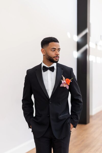 Wedding Photographers in RI | Kelsey Sheehan Photography Timeless Rhode Island Weddings | A man in a black tuxedo with a bow tie and an orange boutonniere stands confidently in a bright, modern room, gazing to the side with a calm demeanor.