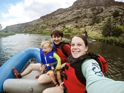 A fun and adventurous family moment captured by an Albuquerque family photographer. This image features a family selfie while canoeing on the river, with everyone wearing red life jackets. The man carries a baby as they sit together in a blue and gray canoe, showcasing a joyful and memorable experience in nature.