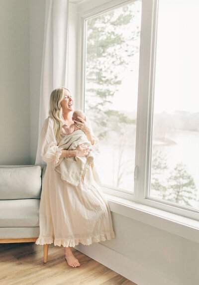 A mother in a cream dress stands barefoot by a large window, peacefully holding her baby during an in-home Kansas City newborn session.