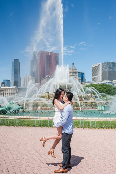 Engaged couple sharing a hug and lift in front of Buckingham fountain and downtown Chicago.