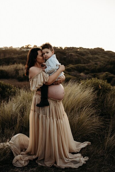A pregnant woman in a flowy dress stands in a field holding a young child as the sun sets behind them, beautifully captured by an Illawarra maternity photographer.