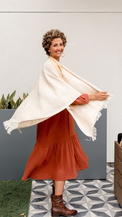 A joyful woman with short curly hair, wearing a flowing rust-colored dress and a cream shawl, twirls with a radiant smile. She exudes confidence and warmth, standing against a modern minimalist backdrop with greenery.