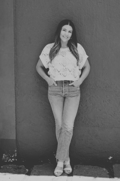 Natalie standing against a terracotta red wall in a cream knit top and jeans, smiling with hands in pockets, ready to connect.