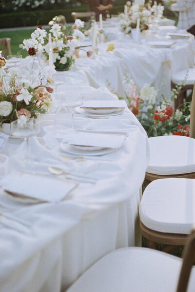 Elegant outdoor wedding table set with white linens, floral centerpieces of white and pink flowers, and place settings