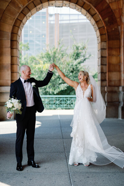 Groom twirls his bride under the historic stone archway at The Pennsylvanian in downtown Pittsburgh, her veil flowing as they laugh together.