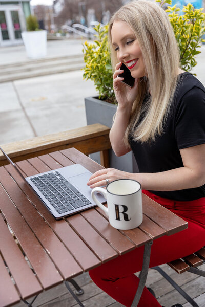 Kristyn sitting in an outdoor cace on her phone with her laptop and a cup of coffee