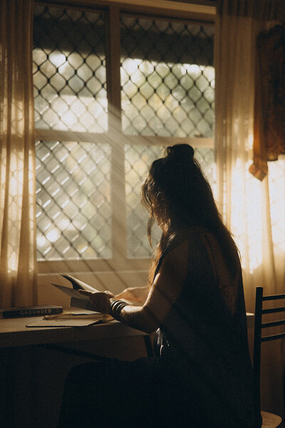 Cinematic personal branding photography featuring a woman reading at a sunlit desk, with golden light streaming through the window. Storytelling brand photography for creatives and entrepreneurs, captured on the Sunshine Coast.