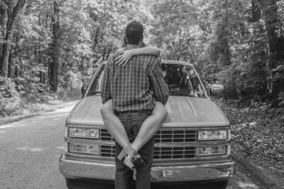 Black and white photo of woman on hood of truck wrapping her legs around a man embracing her