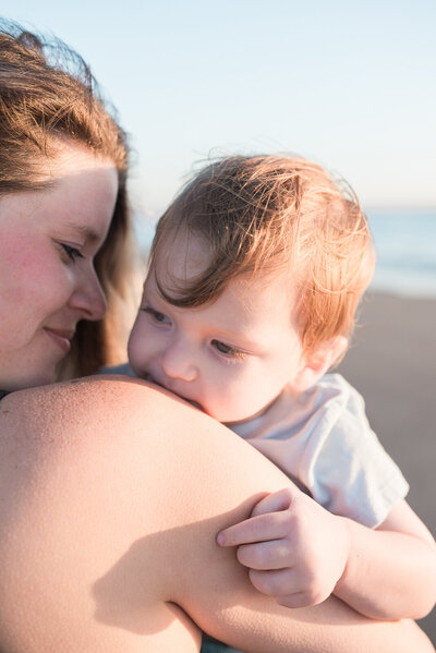 Mom with long hair wearing a black tank top dress, holding her son with his legs wrapped around her waist, spinning him around at sunset during their beach family photos