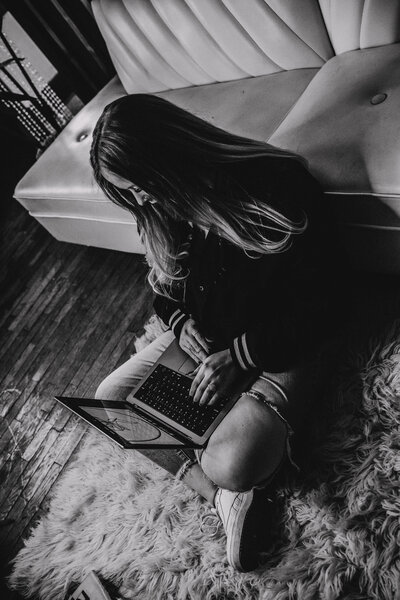 A black and white photo of a woman with long hair sitting on the floor looking at a laptop.