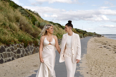 Two brides, hand in hand, stroll along the sandy shore in a touching moment captured by Carry Your Heart Photography. Against the backdrop of the ocean's gentle waves and golden sunset hues, their love shines brightly, symbolizing the beauty and strength of their union on this special day.