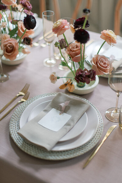 A wedding table setting with gold silverware, roses, soft pink wine glasses, sage green napkins, and white plates