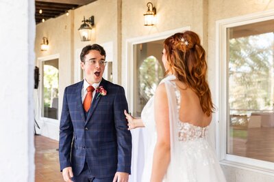 A groom in a navy plaid suit with an orange tie reacts with wide-eyed excitement as he sees his bride for the first time at Tennessee River Place. The bride, wearing a delicate lace gown with flowing sheer sleeves, touches his arm, her auburn curls pinned with star-shaped hair accessories, as sunlight streams through the venue's covered patio