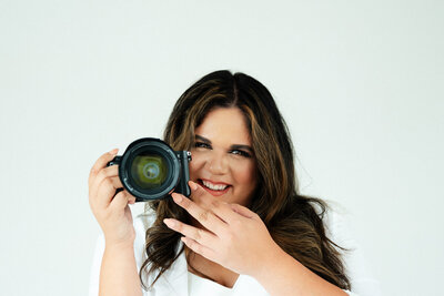 Adriana holding her camera in her right hand with it aimed at the camera taking her photo. She is smiling and partially covering a small part of her face with the camera.