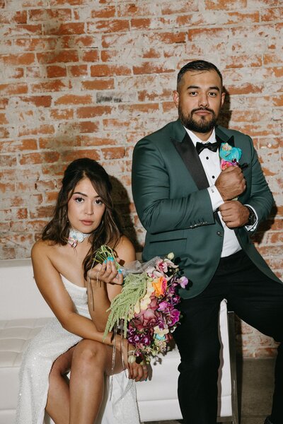 A bride and groom sit on a white sofa, the bride holds a colorful bouquet and the groom adjusts his sleeve