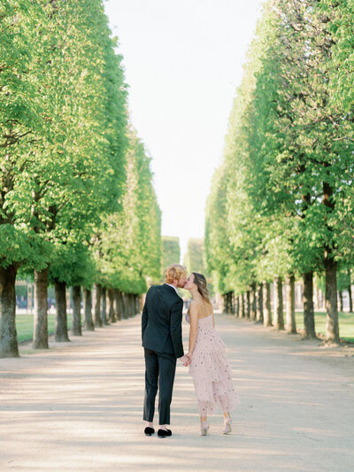 a couple walking away from the camera in a row of trees kissing at jardin du luxembourg