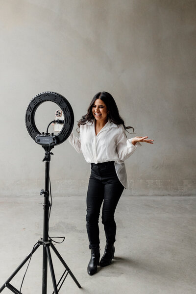 A business and Instagram coach standing confidently in front of a ring light during a personal branding photoshoot in a minimalist studio, showcasing her vibrant and approachable personality.