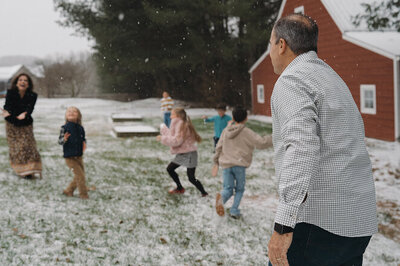 A family with a mom, dad, and five kids are all running around outside in the snow. Behind them is a red barn.