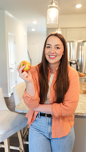 A dietitian in a kitchen holds an apple in her hand, smiling as she prepares to cook.
