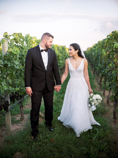 Groom wearing navy blue suit from Charles Tyrwhitt and bride wearing dress from BHLDN hold hands as the sun is setting in the vineyard at RGNY after their wedding ceremony.
