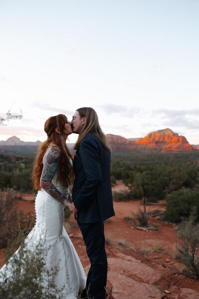 A couple standing on a small bridge, with the woman leaning against the railing, arms splayed, while the man stands behind her, gently kissing her cheek, creating a tender, romantic moment.