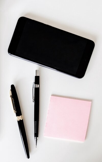 two black pens laying next to each other near a stack of pink post-it notes and a cellphone