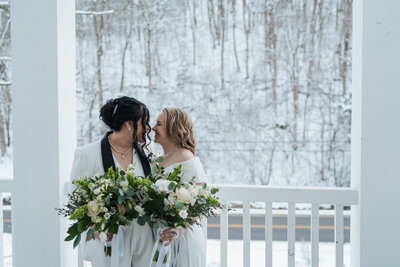Two women stand nose to nose on a porch holding their white wedding bouquets in hand 