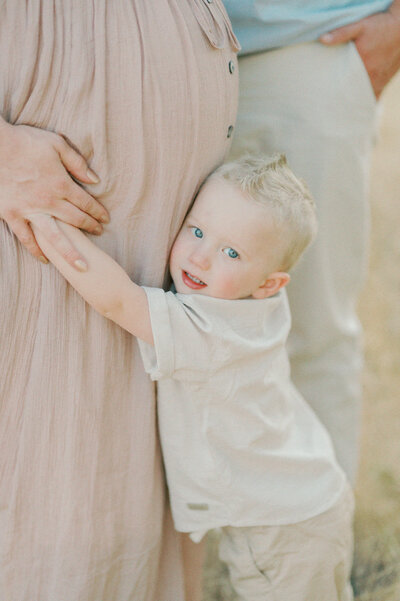 young son with big blue eyes hugging moms belly in a field