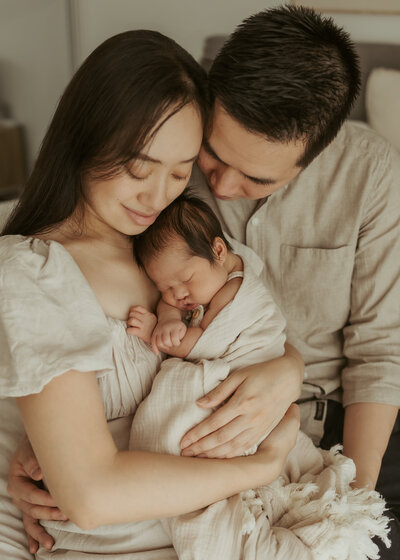 A family of three dressed in neutrals with the newborn sleeping baby curled up against them for their first photoshoot together in their home.