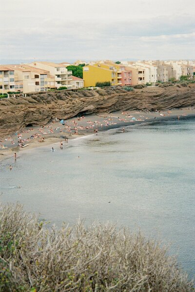 Beach filled with people relaxing near rocky cliffs and colorful coastal apartments under a cloudy sky.