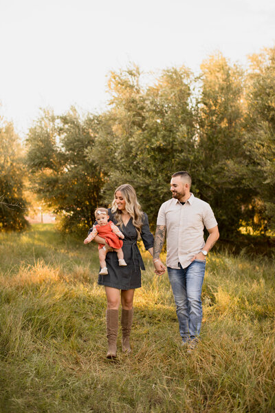 Image of a family of three walking in a green grassy olive tree orchard while looking and smiling at eachother