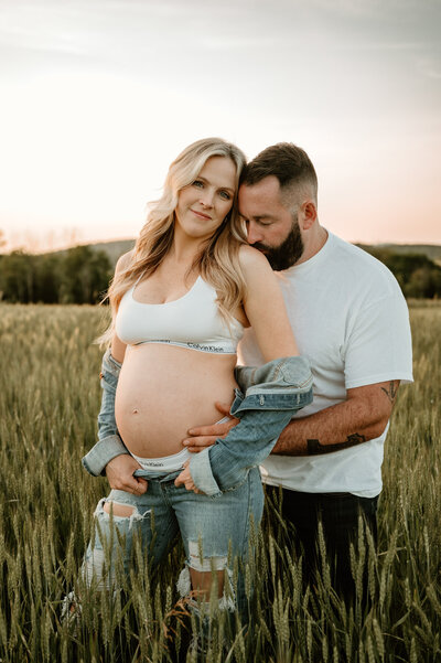 Husband wraps his arms around his expecting wife while they stand in a lush green field. 