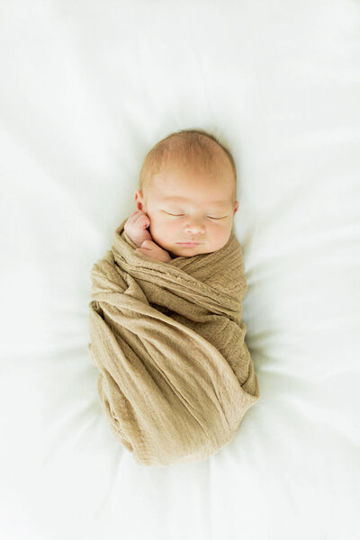 new baby sleeps while swaddled in a tan blanket while laying on a white bed