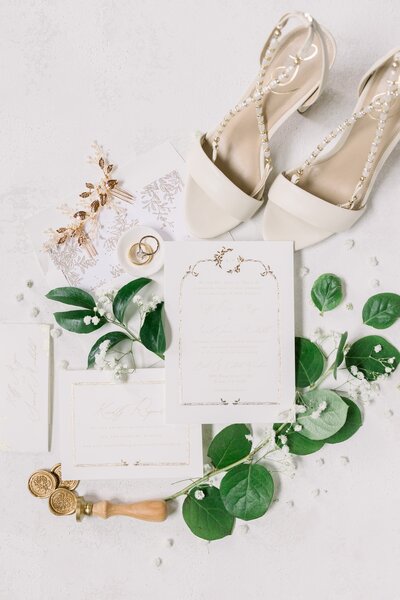 flat lay of wedding details including green leaves, cream sandal high-heels, and invitations