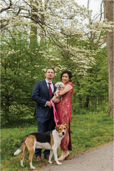 Couple in a red dress and blue suit standing outdoors with their two dogs.