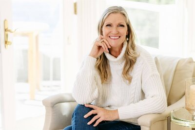 A woman with long blonde hair, wearing a dark blouse and jeans, sits smiling on a bed with patterned pillows in the background—capturing the relaxed spirit of Sweet Pea Ink Creative’s approach to web design.