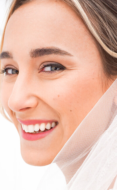 A bride grins happily into the camera. Her beauty is radiant and soft. She wears a peachy pink lip for her wedding day.