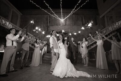 A bride and groom stand smiling under string lights at night, surrounded by their wedding party holding sparklers in celebration. Everyone is dressed formally and appears joyful.