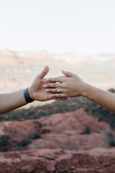 Couple holding hands with diamond engagement ring in focus with mountains in background at Cathedral Rock in Arizona.