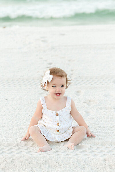 A baby girl with a white bow smiles sweetly while sitting in the soft white sand, dressed in a floral romper by the shoreline.