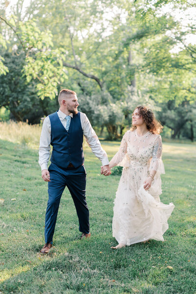 Golden-hour engagement portrait in a green field; embroidered dress and navy waistcoat, candid smiles.
