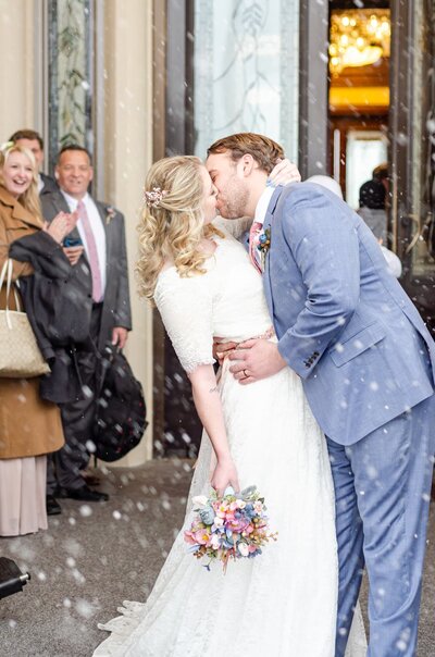 A bride and groom kiss as snow falls around them