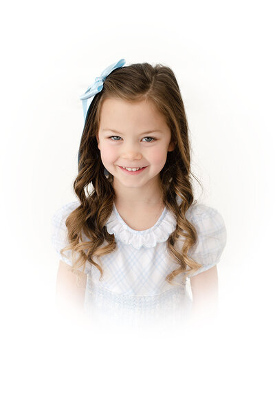 A young child with curly brown hair and a large bow in her hair, wearing a blue and white plaid dress, gazes up at the camera. This charming portrait captures the innocence of childhood against a clean white background, showcasing the art of photography.