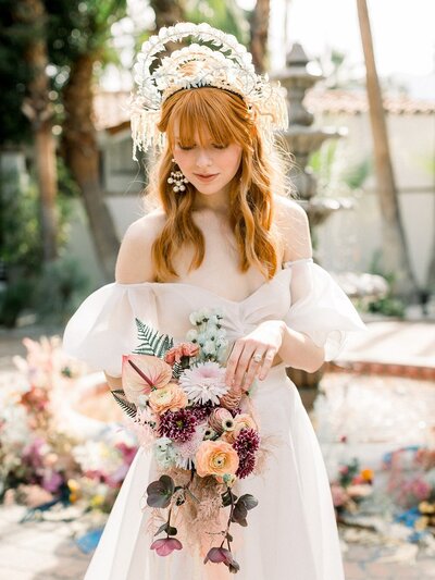 Kohnur bohemian high-fashion bride with natural subtle makeup and large fringe crown, wearing off-shoulder gown in a Palm Springs villa courtyard.