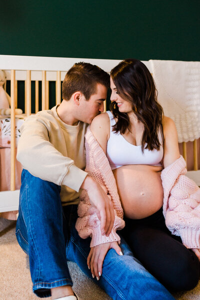 A photo of a husband and wife in their nursery with a crib behind them. The wife is very pregnant and has er belly exposed. The husband is kissing her shoulder.