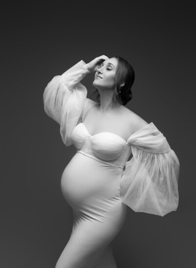 High-fashion maternity portrait of a woman in a sheer puff-sleeve neutral gown with dramatic lighting and a classic pose