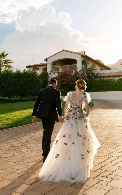 A bride and groom walk away from the camera holding hands, up a stone driveway to a spanish-looking manor. The grooms suit is black and the wedding dress is toile decorated with colourful flower appliques.