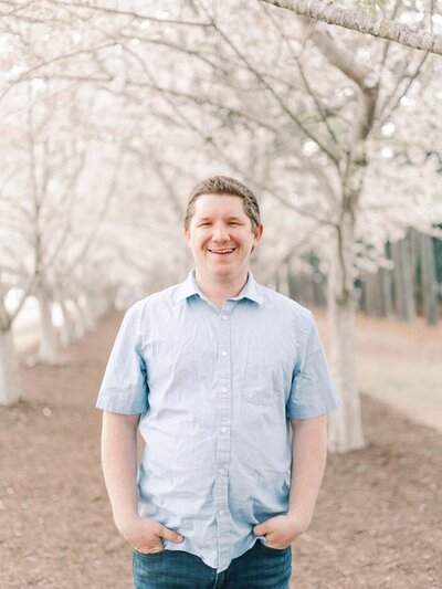 White man stands between white cherry blossom trees smiling, wearing a blue button up shirt.
