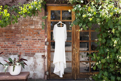 White wedding dress hangs up in doorframe outside of brick, greenery covered building